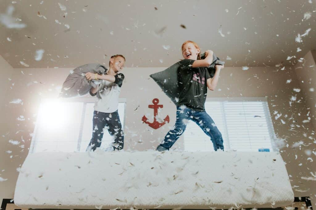 Two young children having a pillow fight on a bed with feathers falling all around them