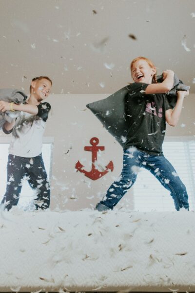 Two young children having a pillow fight on a bed with feathers falling all around them