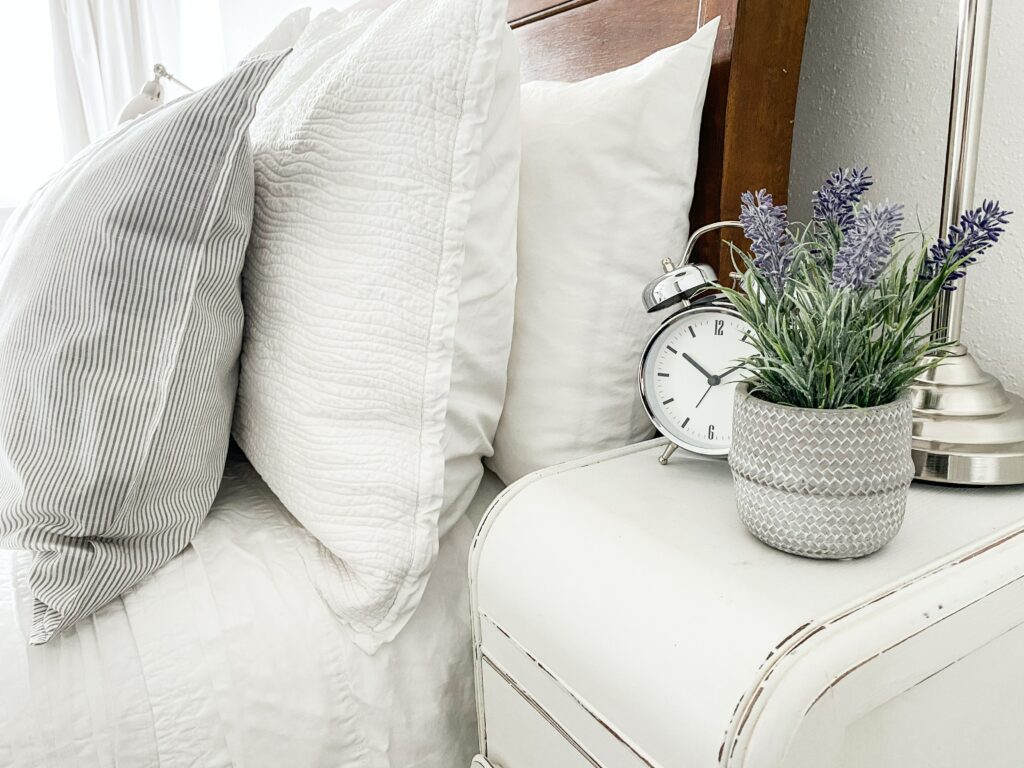 A small silver alarm clock sits next to a lavender plant on a white nightstand next to a bed with white bedding