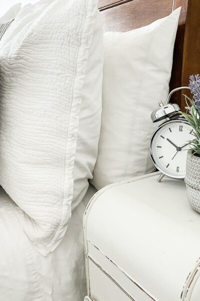 A small silver alarm clock sits next to a lavender plant on a white nightstand next to a bed with white bedding