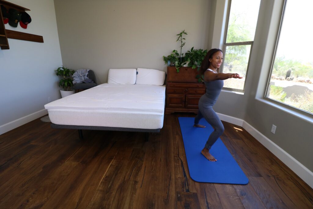 A woman doing a yoga pose next to a bed with a Naturally Nestled latex mattress topper and pillows