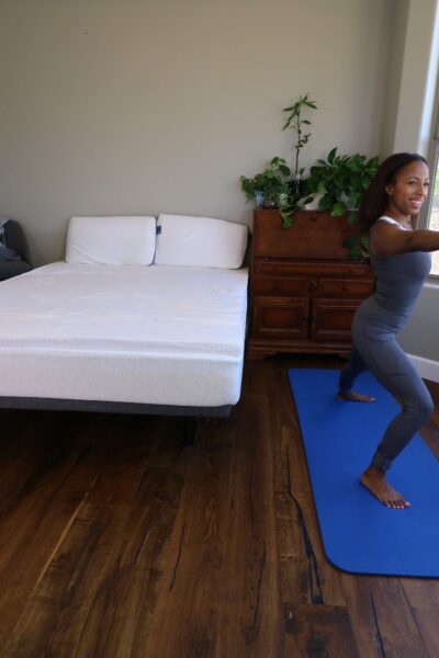 A woman doing a yoga pose next to a bed with a Naturally Nestled latex mattress topper and pillows