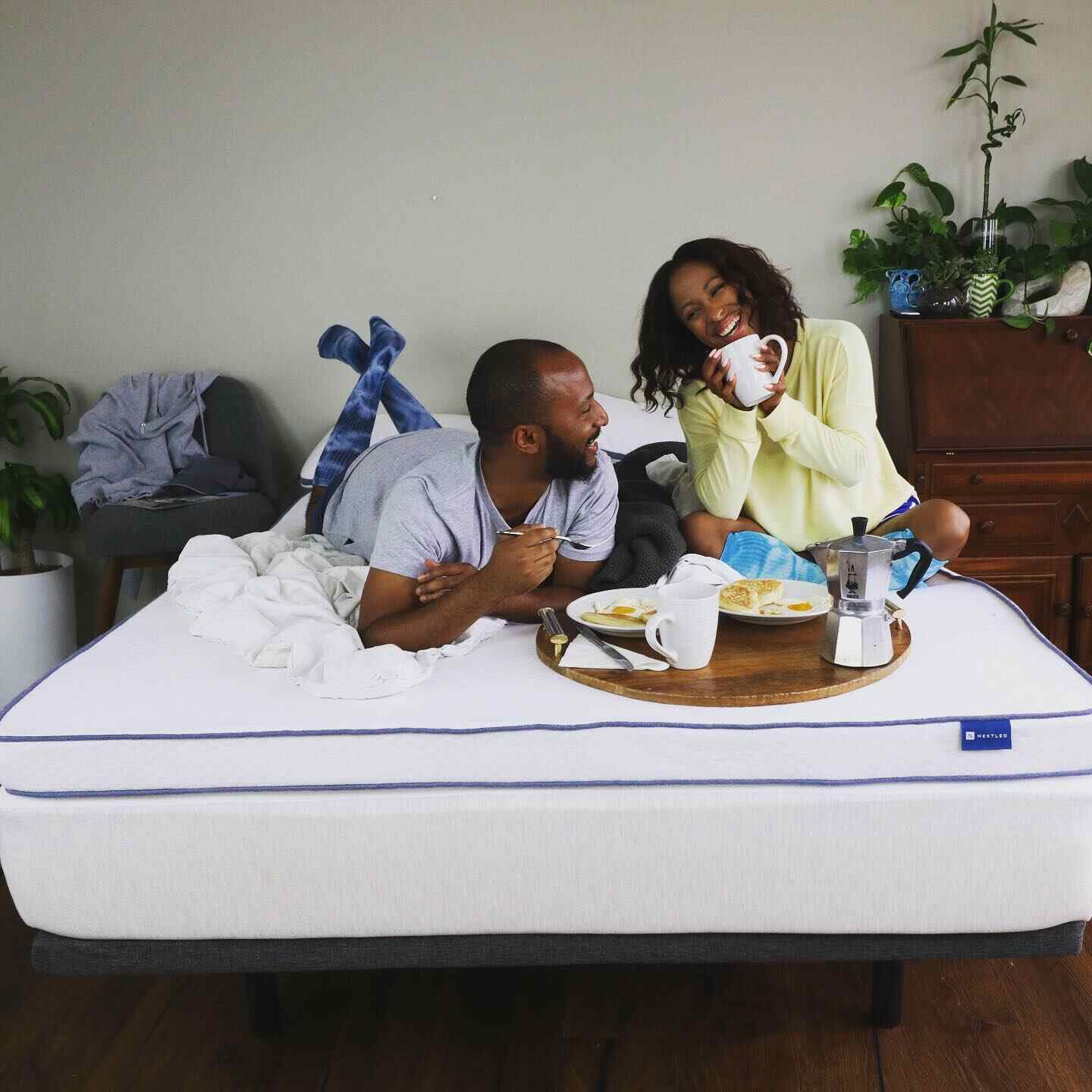 A man and a woman sit on an eco-friendly mattress topper and eat breakfast on a tray.