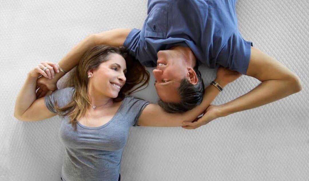A man and woman smile at each other while laying on an eco-friendly mattress topper.