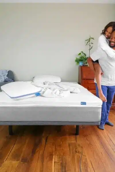 A man with a woman on his back stands next to a Naturally Nestled mattress and smiles