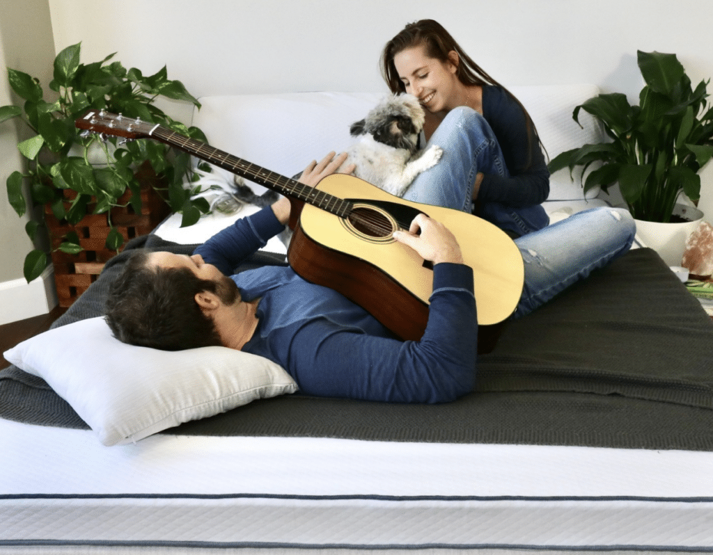 A happy couple is on a sustainable latex mattress playing with a guitar and a dog