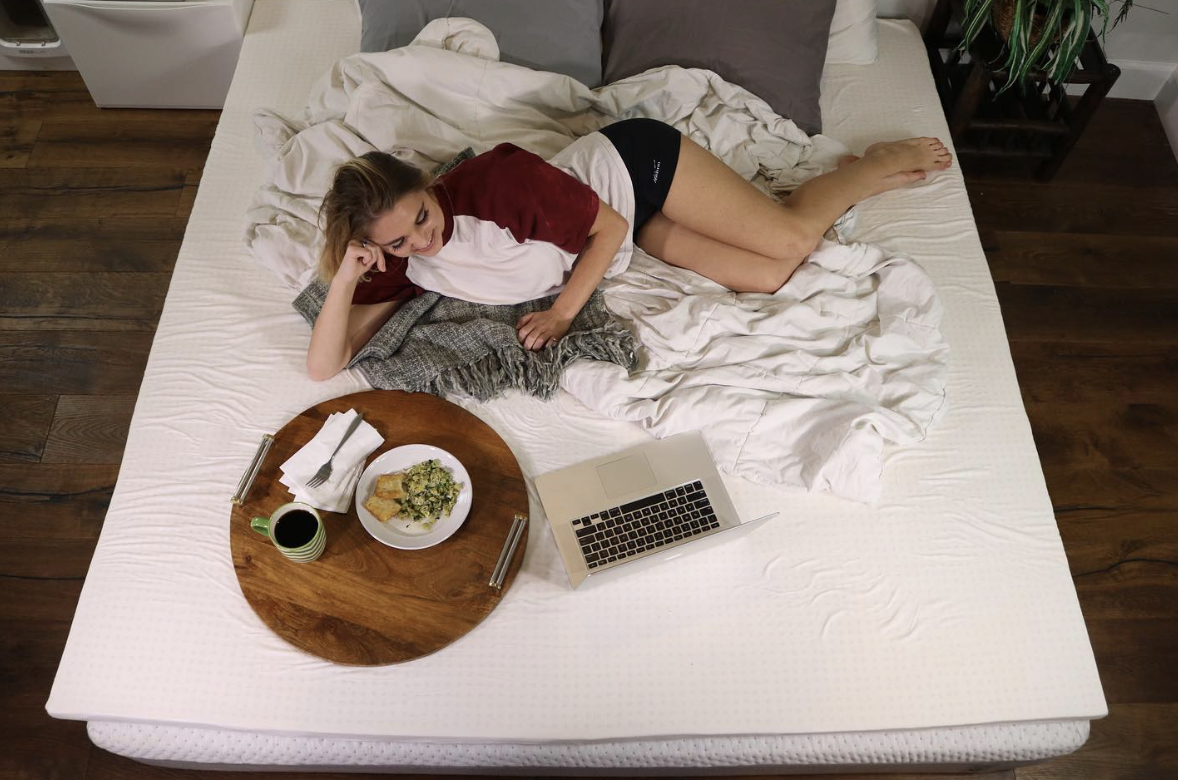 A woman watches a show while eating food and relaxing on a Naturally Nestled mattress free of fiberglass