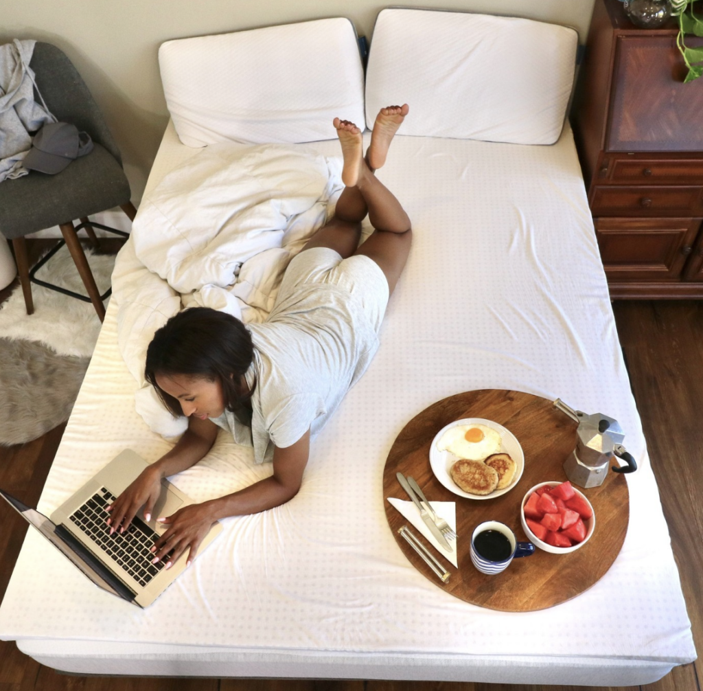 Young woman lays on her firm mattress topper, working on a laptop with breakfast next to her